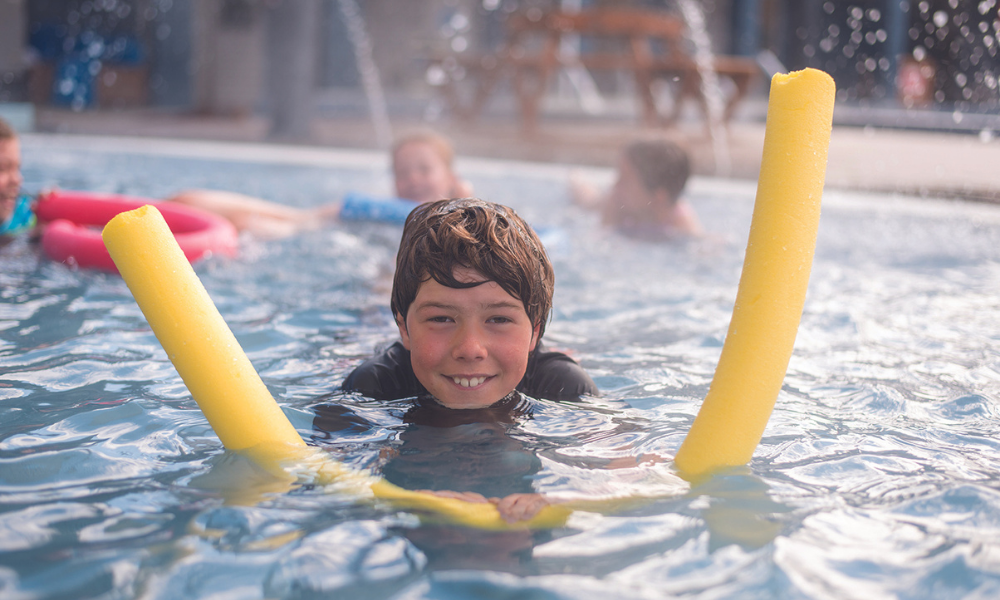 Boy using swimming noodle in pool smiling at camera